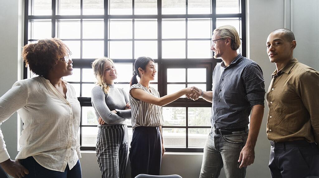 Vijf mensen in een vergaderruimte, twee schudden handen.