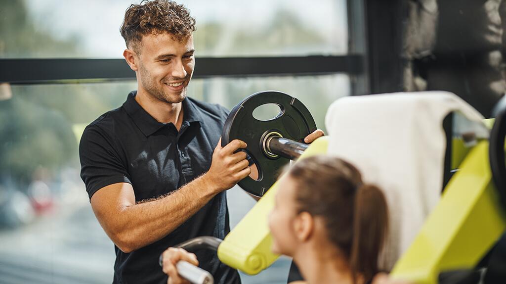 Man helpt vrouw met gewichtheffen in de sportschool, beiden glimlachend.