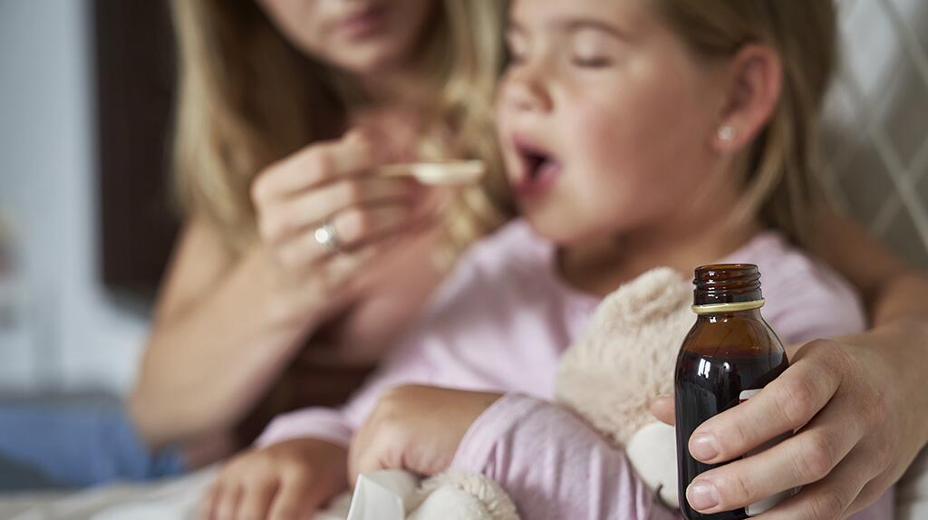 Moeder geeft zieke dochter medicijnen, knuffelbeer in hand.
