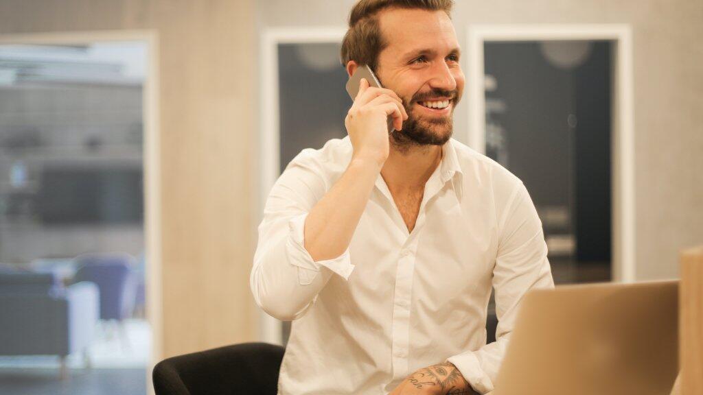 Man in witte blouse glimlacht terwijl hij belt aan een bureau met laptop.