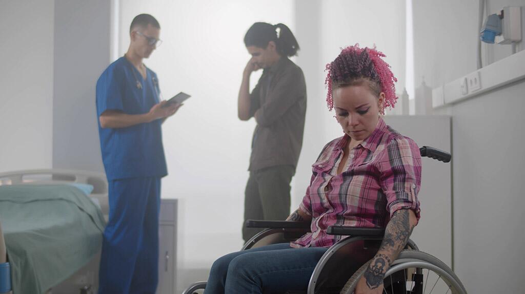 Vrouw in rolstoel met roze haar, arts in blauw uniform, persoon met handen voor gezicht.