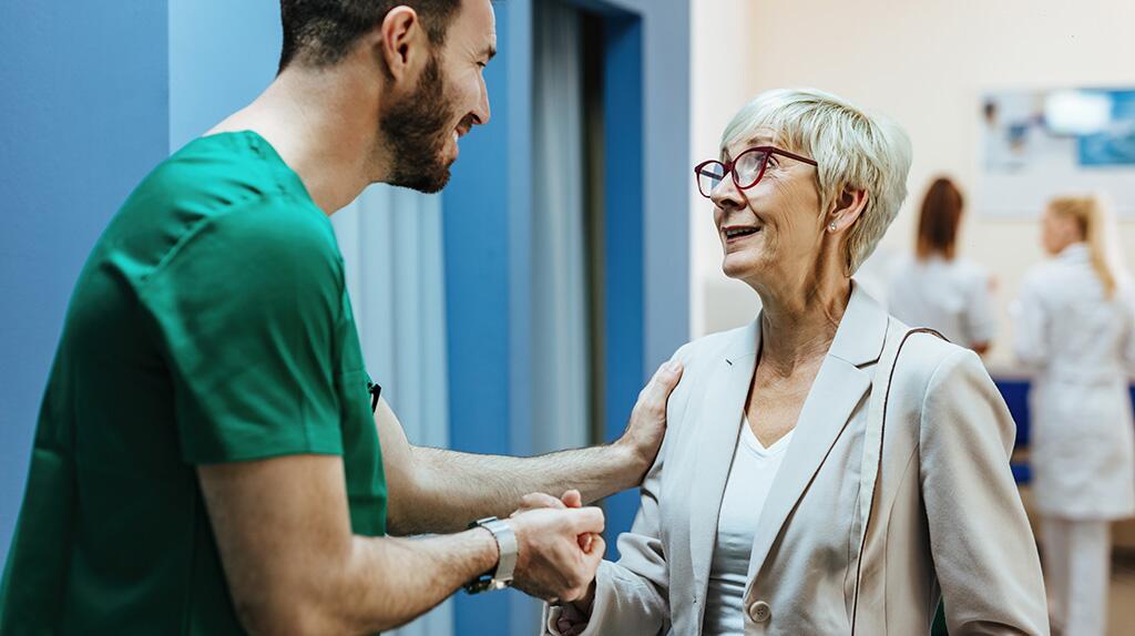 Verpleegkundige en oudere vrouw glimlachen in een ziekenhuisgang.