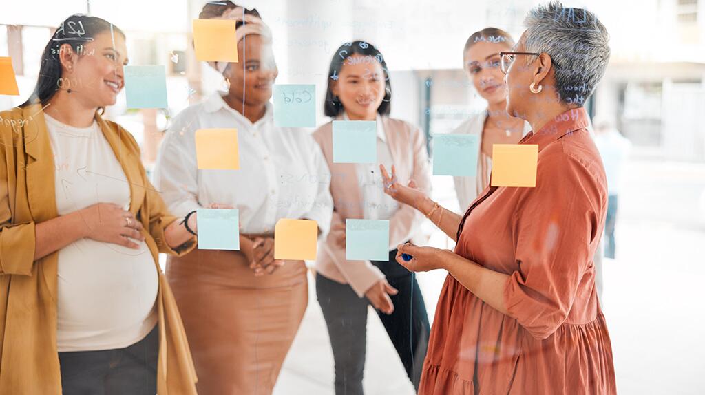 Vijf vrouwen bespreken notities op een glazen muur.