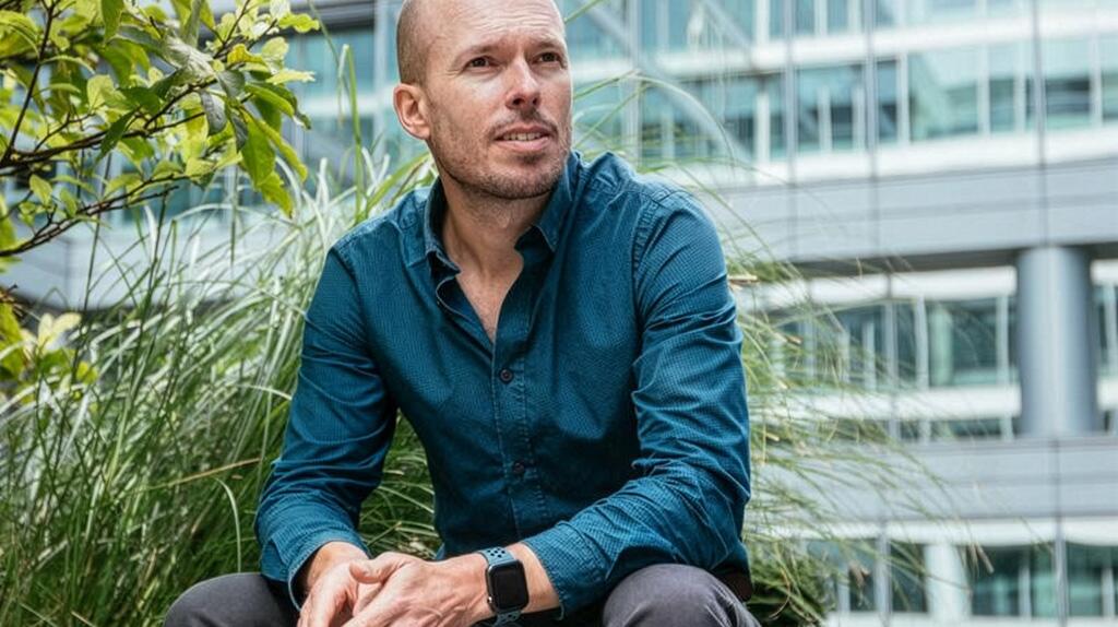 Man in blauwe blouse zit buiten, kijkt opzij, moderne achtergrond.