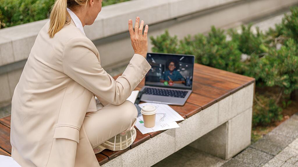 Persoon in beige pak videobelt buiten op laptop, met koffie en papieren naast zich.
