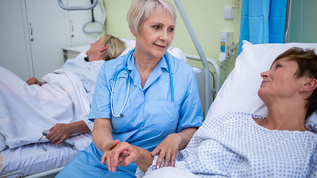 Zuster praat glimlachend met patiënt in ziekenhuisbed.
