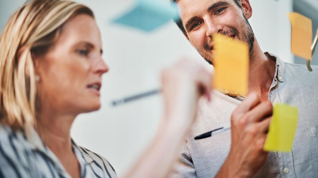 Twee mensen brainstormen met gekleurde post-its op een muur.