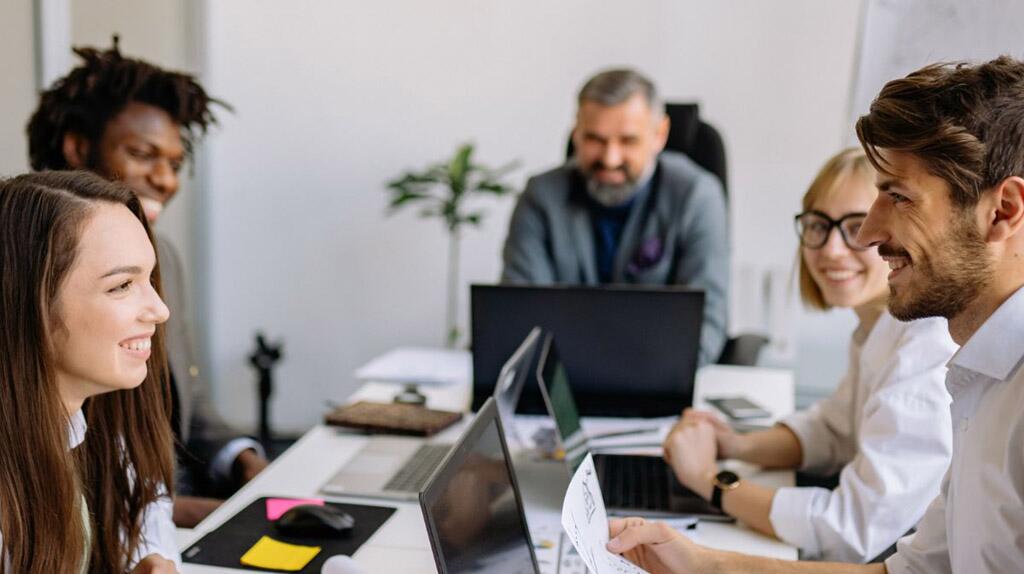 Collega's lachen tijdens een vergadering rond een tafel met laptops.