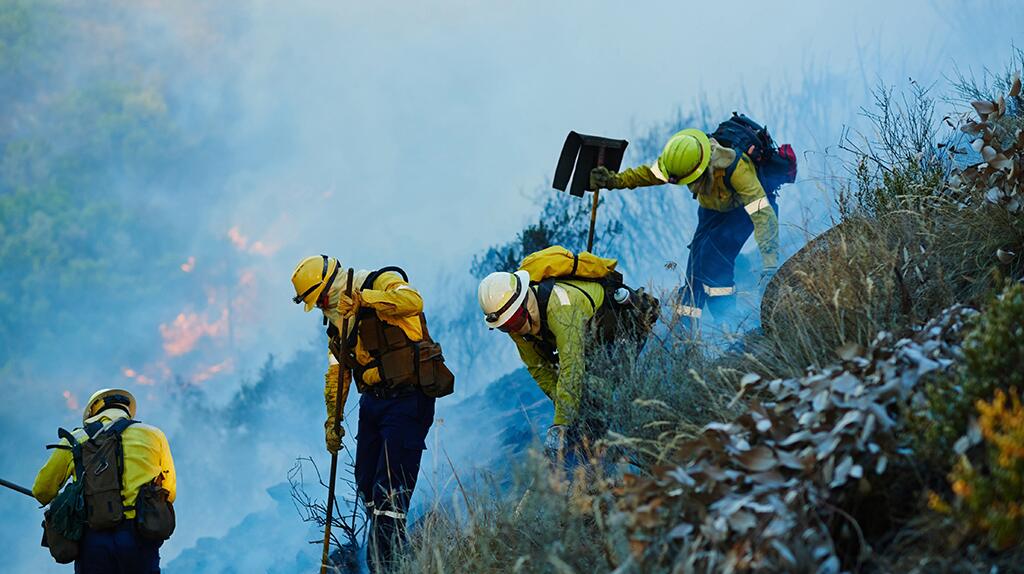 Brandweerlieden blussen vuur in rookachtige heuvels, dragen gele helmen.