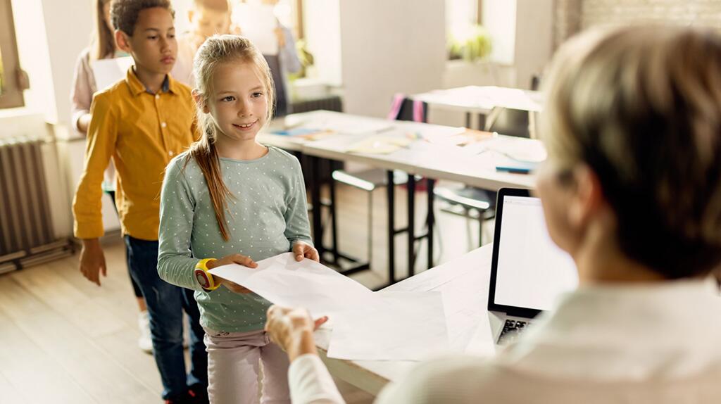 Kinderen geven documenten aan een persoon achter een bureau in een klaslokaal.