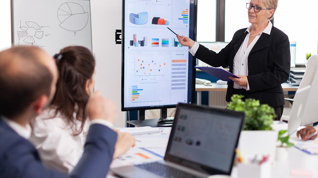 Vrouw geeft presentatie aan collega's met scherm in kantooromgeving.