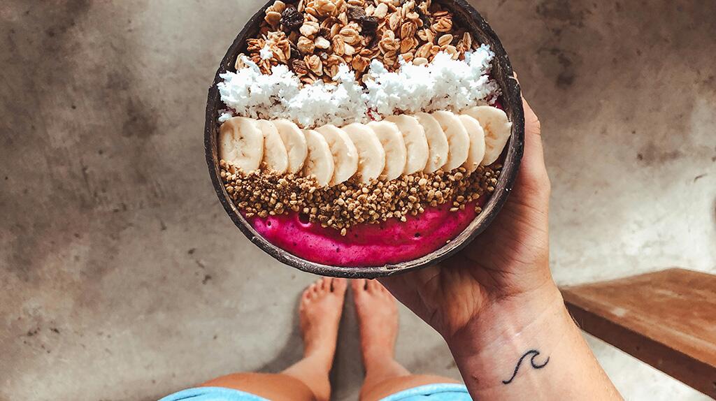 Smoothiebowl met banaan, walnoten, kokos en pitten; hand met polstattoo houdt de kom vast.