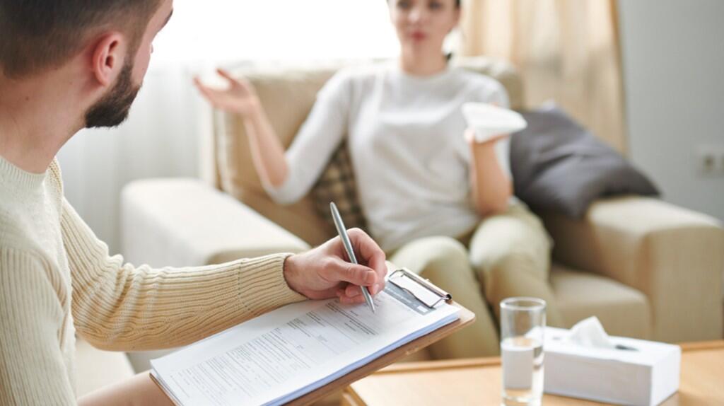 Man maakt notities, vrouw praat op bank, glas water op tafel.