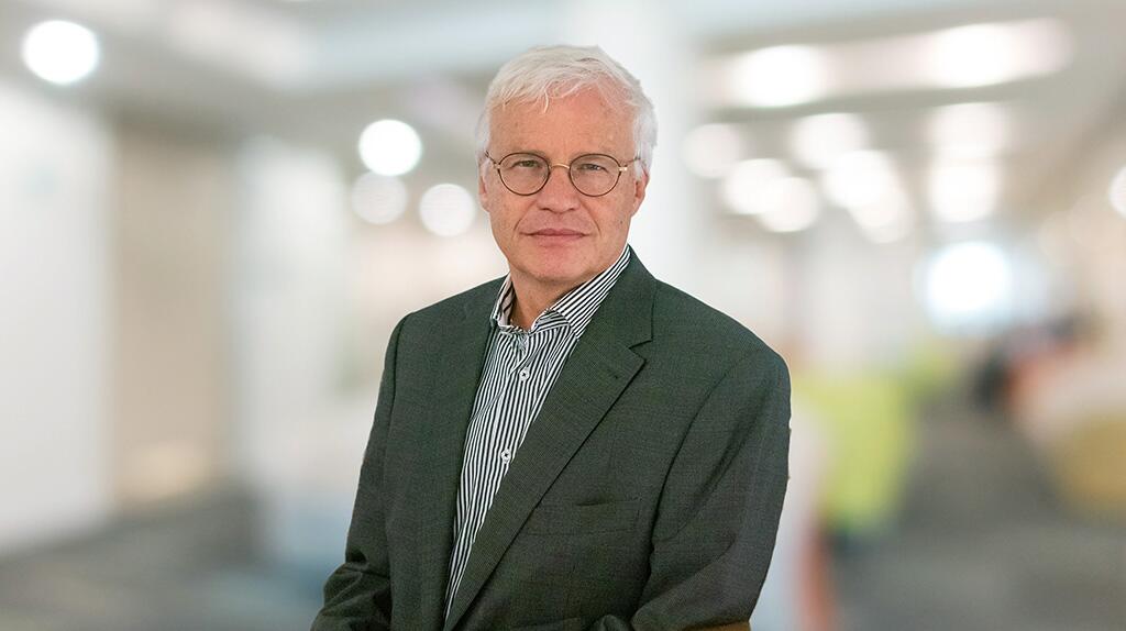 Oudere man in pak glimlacht, onscherpe kantooromgeving op de achtergrond.