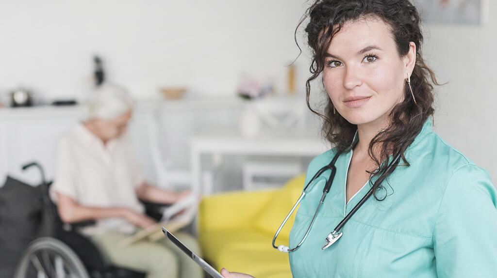 Vrouwelijke arts in groene jas met stethoscoop, oudere vrouw op achtergrond in rolstoel.