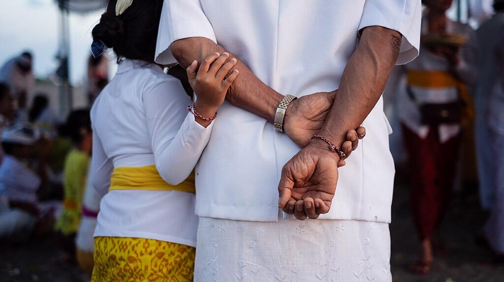Persoon in witte kleding met kind dat hem omarmt, kleurrijke kleding op de achtergrond.