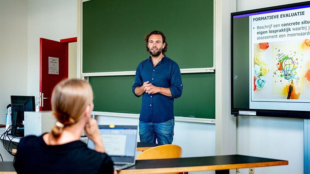 Man presenteert in klaslokaal, laptop en projectiescherm zichtbaar.