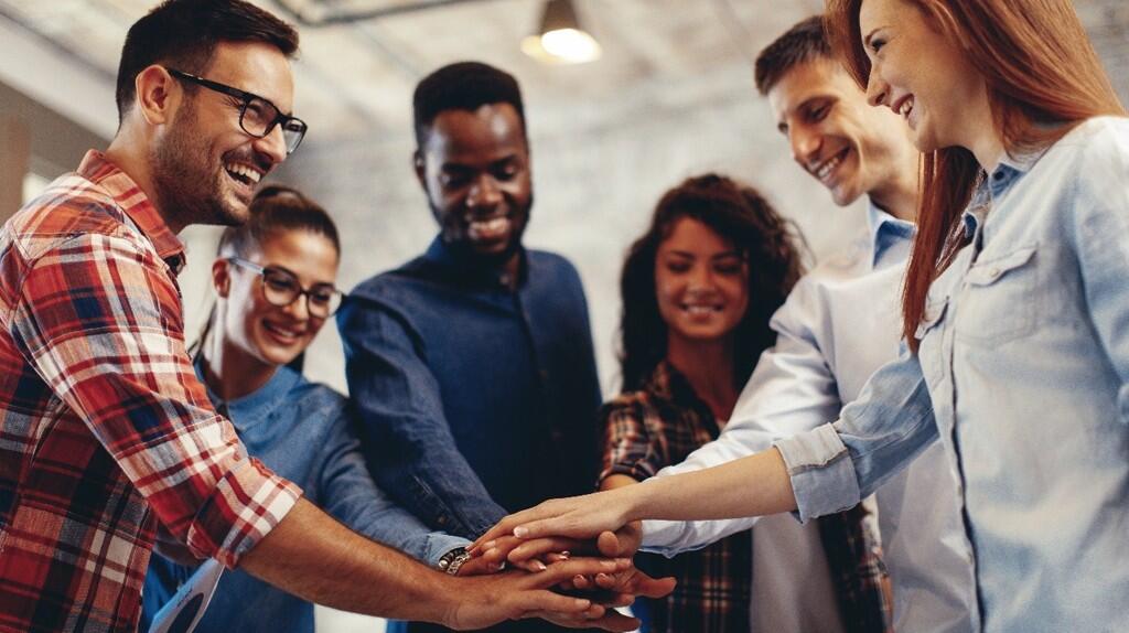 Mensen in een kring leggen hun handen op elkaar, glimlachend en samenwerkend.