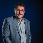 Man met bril en snor in blauw pak tegen donkere achtergrond.