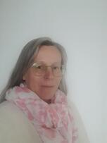 Vrouw met grijs haar en bril draagt een roze sjaal en witte trui, staat voor een witte muur.