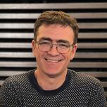 Man met bril glimlacht, draagt een donkere trui. Houten achtergrond.