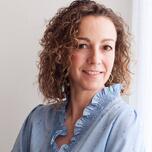 Vrouw met krullend haar in lichtblauwe blouse, glimlachend voor neutrale achtergrond.