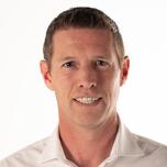 Een glimlachende man met kort bruin haar in een wit overhemd, tegen een lichte achtergrond.