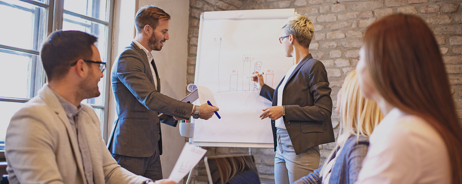 Zakelijke vergadering met vijf personen bij een flip-over, focus op samenwerking.