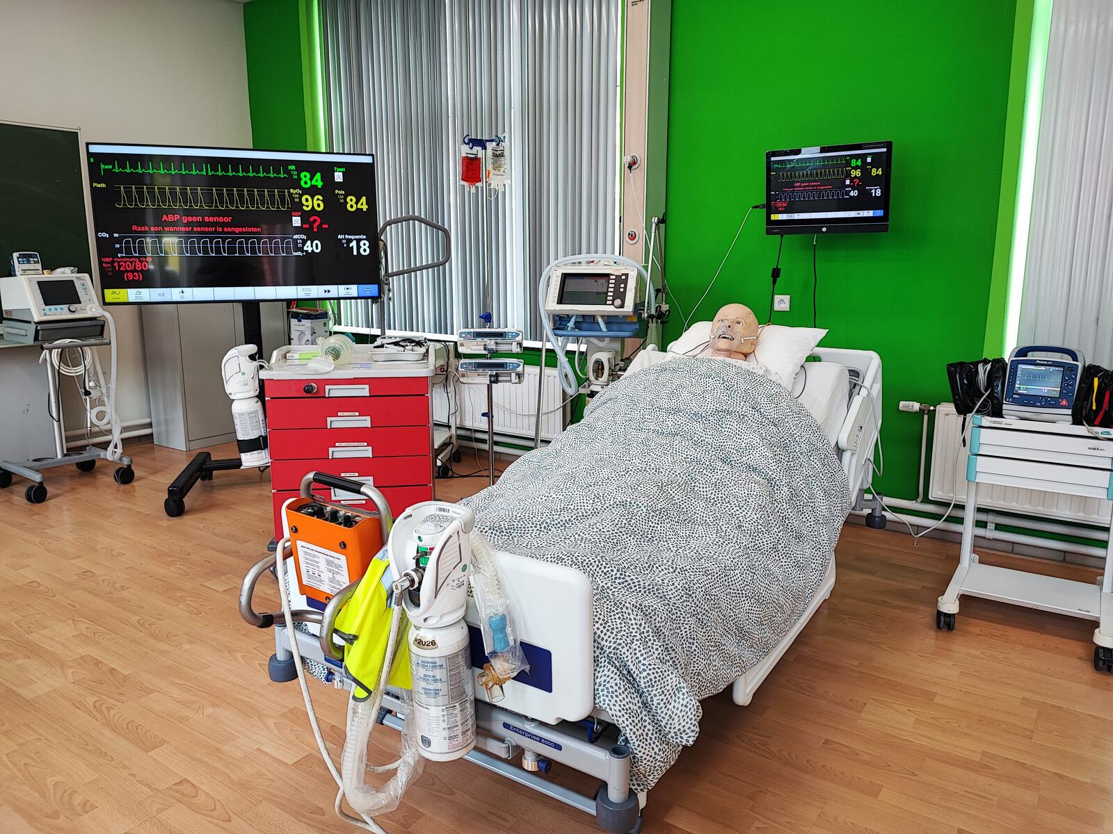 Ziekenhuiskamer met apparatuur en een ziekenhuisbed met een pop onder een grijze deken.