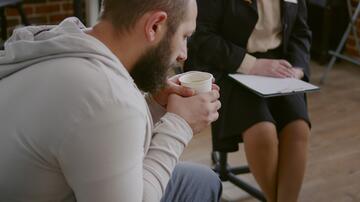 Man met kopje in gesprek met vrouw op kantoorstoel, papier in haar handen.
