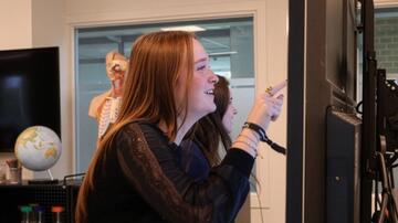 Vrouw lacht naar scherm in kantoorruimte. Globes en apparatuur op achtergrond.