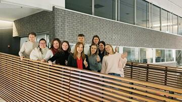 Groep mensen lacht en zwaait op een moderne binnenlocatie met houten balustrade.