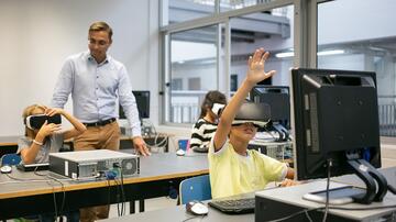 Kinderen met VR-brillen in klas, jongen steekt hand op, leraar kijkt toe.