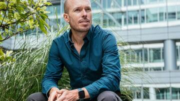 Man in blauwe blouse zit buiten voor modern gebouw en begroeiing.