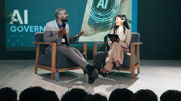 Twee mensen zitten op een podium met een scherm en praten voor een publiek.