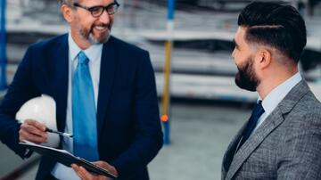 Twee mannen in pakken praten en glimlachen in een industrieel magazijn.