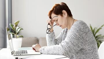 Vrouw met bril leunt vermoeid op hand, zittend bij een laptop.