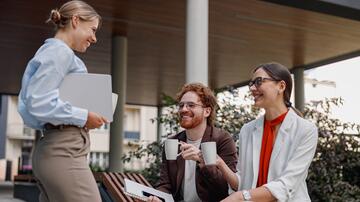 Collega's in gesprek op kantoor met koffie. Vrouw staat, twee zitten lachend op bank.