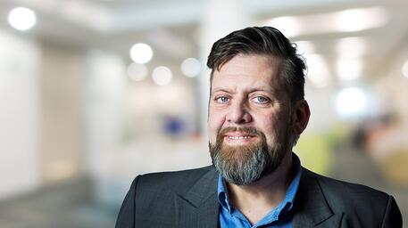 Man met baard in pak, glimlachend in kantooromgeving.