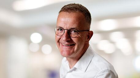 Man met bril in wit overhemd glimlacht, wazige achtergrond.