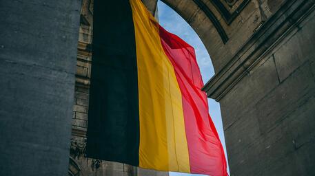 Belgische vlag wappert onder een stenen boog, blauw lucht zichtbaar.