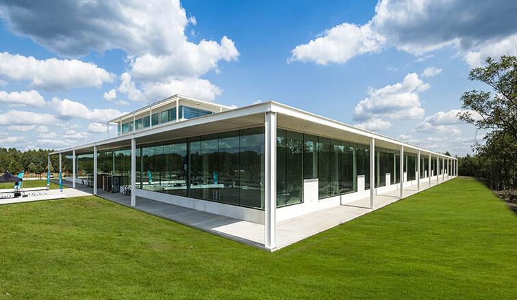 Modern gebouw met glazen wanden op een groene grasvlakte onder een blauwe hemel met wolken.