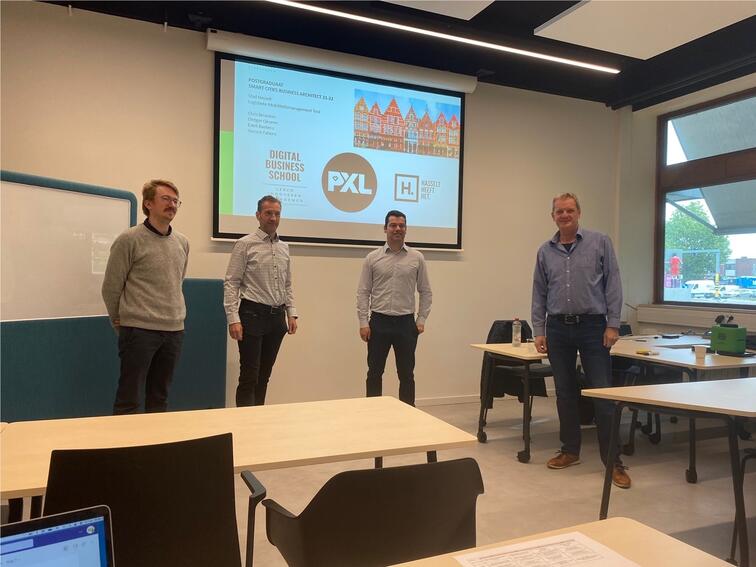 Vier mannen presenteren in een klaslokaal met een projectiescherm.