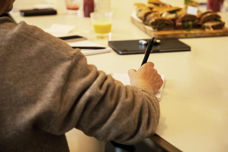 Persoon schrijft aan tafel met notitieblok en pen, eten op de achtergrond.