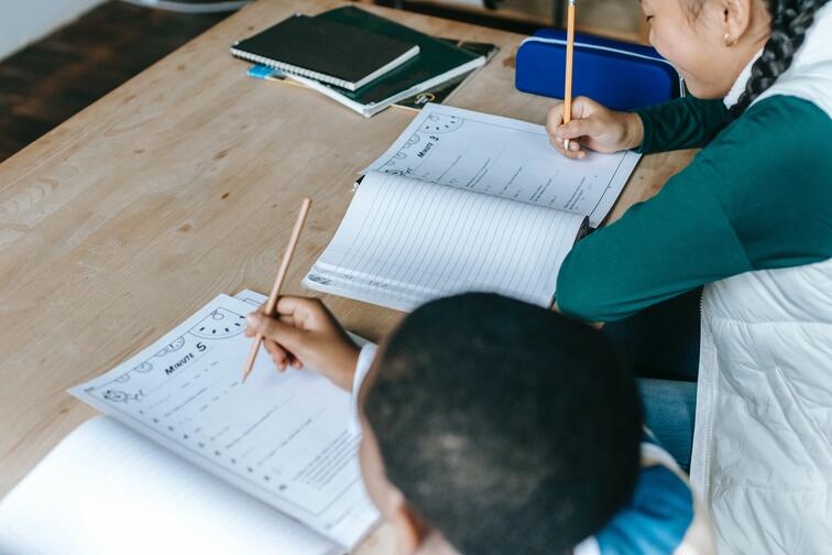 Twee kinderen schrijven in werkboeken aan een houten tafel.