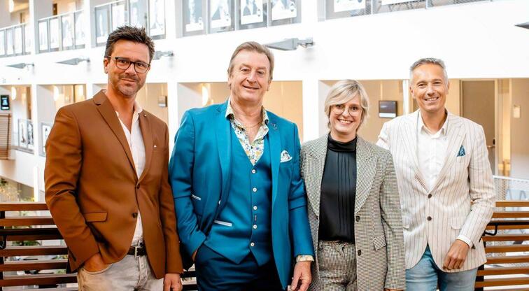 Vier lachende mensen in kleurrijke kostuums in een lichte kantooromgeving.