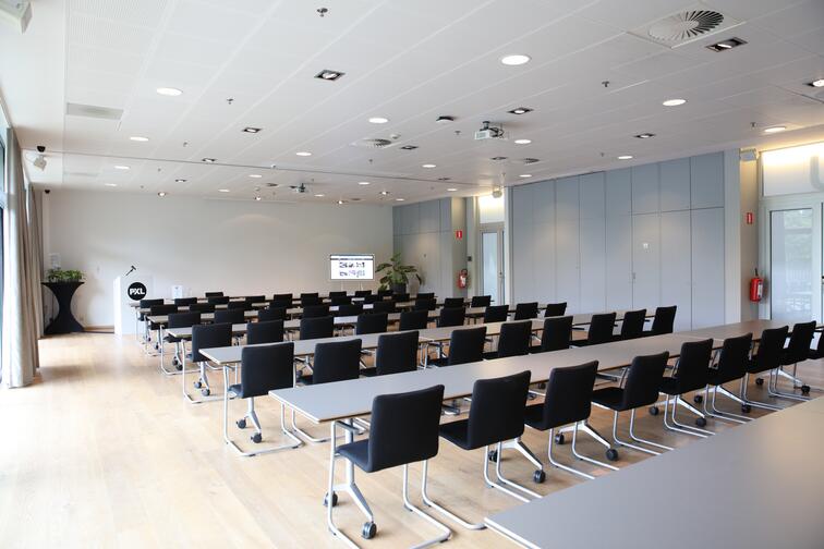 Lege vergaderzaal met zwarte stoelen en tafels, lichte muren en houten vloer.