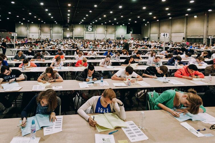 Grote zaal met vele studenten die examen maken aan lange tafels.